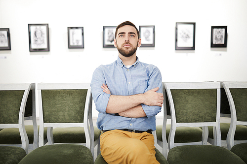 Portrait of pensive bearded young man looking at pictures in art gallery or museum while sitting on velvet chair, copy space