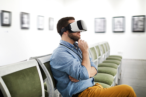 Side view portrait of bearded young man wearing VR while looking at pictures in museum or art gallery, copy space