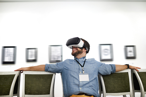 Portrait of smiling bearded young man wearing VR while looking at pictures in museum or art gallery, copy space