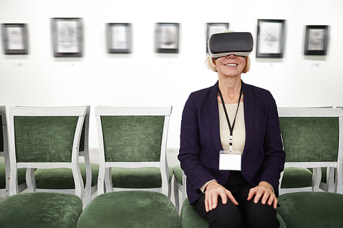 Portrait of smiling mature woman wearing VR while looking at pictures in museum or art gallery, copy space