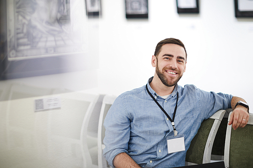 Portrait of smiling bearded man wearing blank name tag sitting on chair in art gallery or museum, copy space