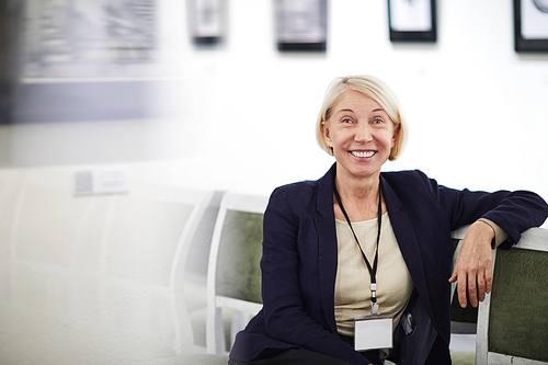 Portrait of smiling mature woman wearing blank name tag sitting on chair in art gallery or museum, copy space