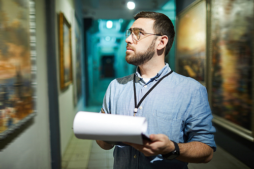 Waist up portrait of bearded man holding clipboard while looking at pictures in art gallery or museum, copy space