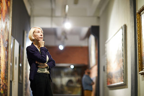 Portrait of pensive mature woman looking at paintings standing in art gallery or museum, copy space