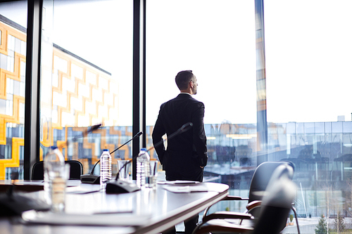 Young businessman or politician in elegant suit standing by window at break in conference hall