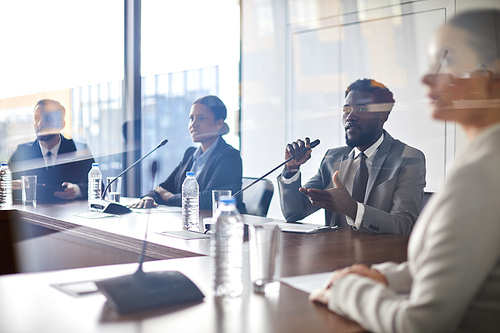 Young African-american delegate in suit speaking in microphone while sitting in conference hall among colleagues