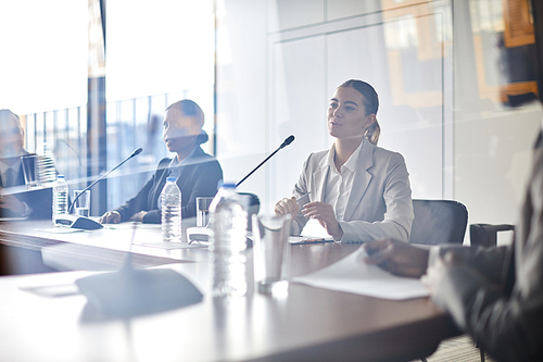 Young elegant businesswoman sitting by table at conference and making report for foreign colleagues