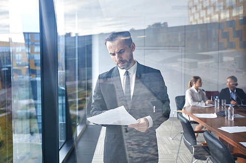 Serious businessman in suit reading text of his report before conference while standing by large window