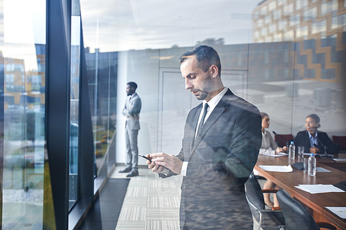 Young serious delegate standing by window and scrolling in his smartphone in conference hall at break