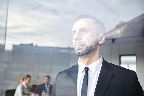 Serious businessman in formalwear looking through window while his colleagues preparing for conference on background