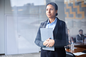 Young serious delegate with clipboard standing by window of conference hall with colleagues on background