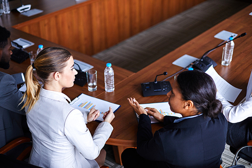 Two young female analysts or economists discussing financial issues before conference while sitting by table