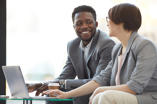 Positive creative multi-ethnic business team sitting in lobby and looking at each other while discussing sales information on laptop