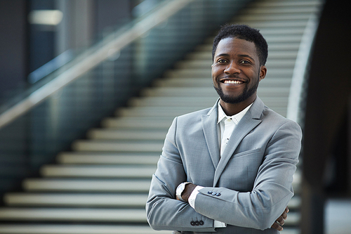 Portrait of jolly successful young black business executive with beard standing against staircase of lobby and crossing arms on chest