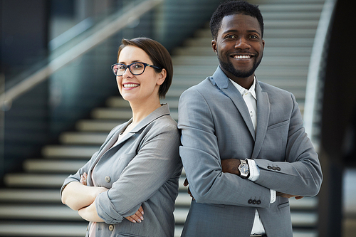 Smiling confident strong multi-ethnic business team members in formalwear standing back to back and keeping arms crossed while 