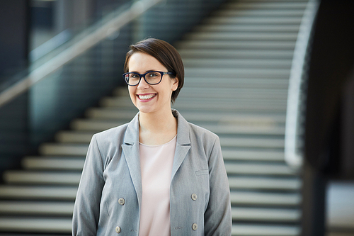 Cheerful excited female project manager with wide smile wearing gray jacket and eyeglasses standing in lobby and 