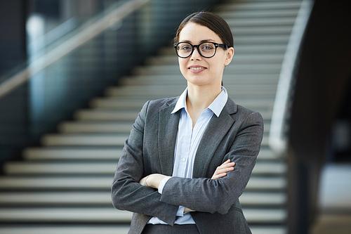 Portrait of positive young female intern in glasses wearing gray jacket standing against stairs and crossing arms on chest