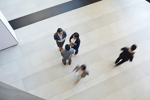 Directly above view of intercultural business colleagues in formalwear standing in lobby of company and chatting during break while another employees being in hurry, blurred motion effect