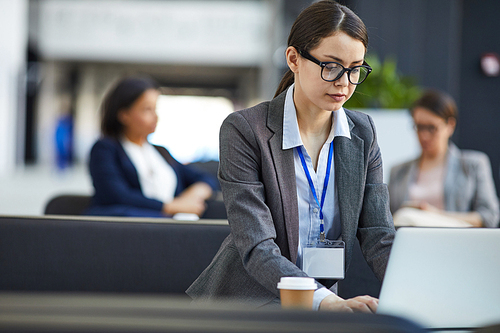 Serious concentrated female conference participant in glasses wearing badge on neck sitting in lobby and checking presentation on laptop