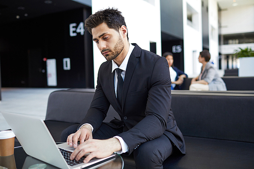 Serious young mixed race manager with beard sitting on sofa in lobby and using laptop while examining data and editing files