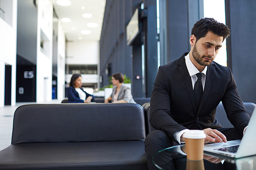 Serious pensive handsome young businessman in black suit sitting in modern hall with sofas and connecting laptop to wifi while working out of office