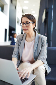 Serious concentrated short-haired lady entrepreneur in glasses sitting on sofa in lobby and using internet on laptop while examining information