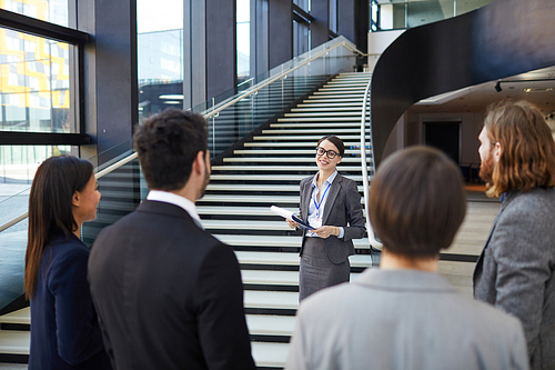 Smiling confident business forum guide with badge on neck standing on stairs and holding clipboards with files while giving tour to participants