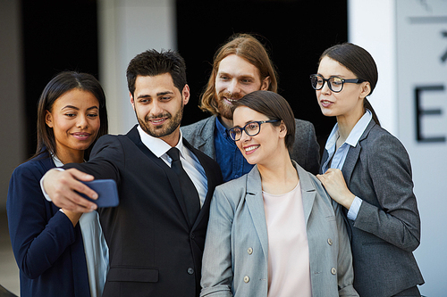 Cheerful purposeful young multi-ethnic business colleagues making group selfie on smartphone in lobby