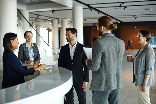 Cheerful young multi-ethnic forum participants in formal outfits standing at registration counter and chatting with receptionist in lobby