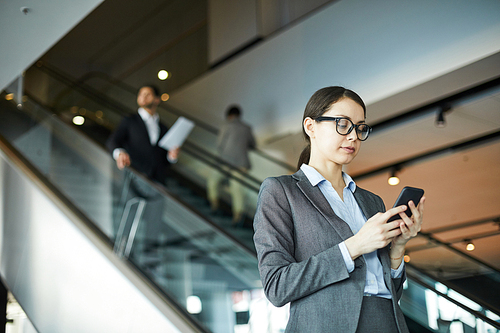 Serious young woman in glasses standing at escalator in business center and checking email on smartphone