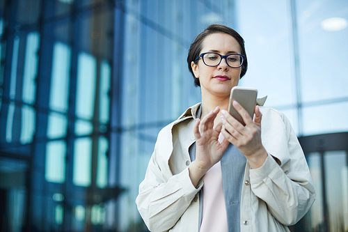 Serious busy attractive middle-aged woman in coat standing against office building and using smartphone while checking email