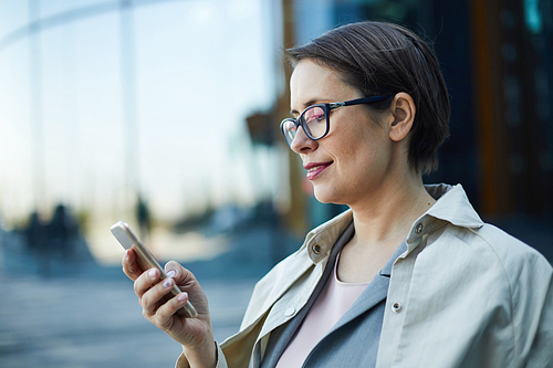 Content attractive business lady with short hair wearing coat and eyeglasses walking over city street and using mobile app on smartphone