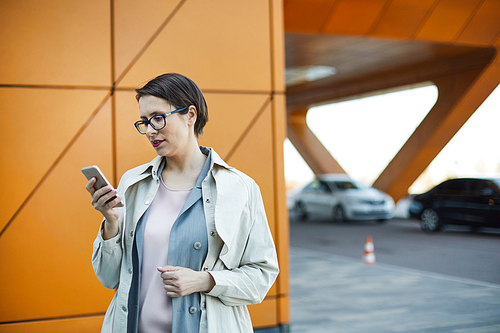 Concentrated attractive middle-aged woman with short hair standing outdoors and using mobile app while ordering taxi online