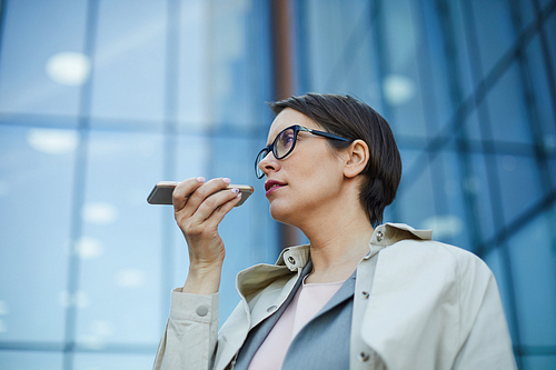 Serious pensive confident middle-aged businesswoman in eyeglasses standing against office building and using smartphone while recording voice message