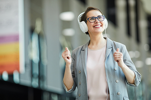 Happy excited middle-aged lady in eyeglasses dancing and looking around while listening to music in headphones