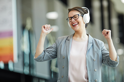 Jolly carefree middle-aged woman in eyeglasses keeping eyes closed and having fun while listening to music in headphones and dancing in excitement