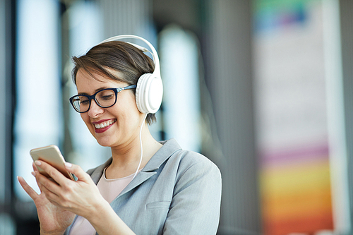 Excited attractive middle-aged woman in wired headphones using modern smartphone while chatting online