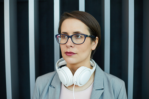 Close-up of serious pensive young businesswoman with headphones on neck wearing eyeglasses contemplating around while being deep in thoughts