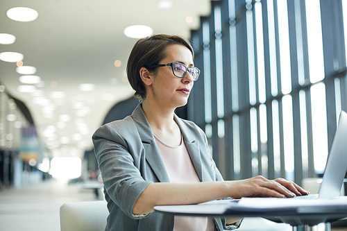 Content enterprising attractive business lady in glasses standing at high table and typing on laptop while working in lobby