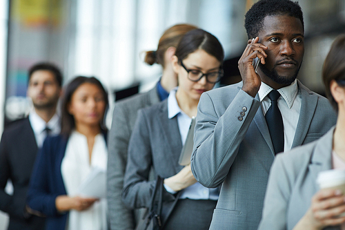 Serious displeased young African-American businessman with beard standing in line and calling on mobile phone while waiting for business forum registration