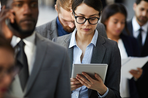 Serious busy attractive young lady in glasses reading document on tablet while standing in long line