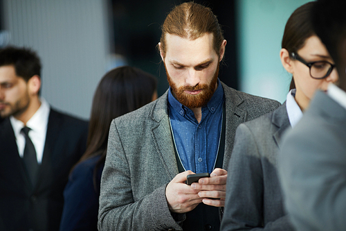 Serious thoughtful young businessman with beard using telephone while checking message and waiting in line