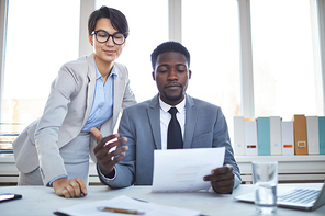 Two young confident intercultural employees reading financial paper by workplace at working meeting