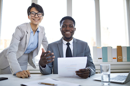 Two young happy colleagues in formalwear looking at you with smiles by desk on background of window