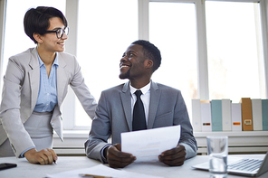 Two cheerful employees in elegant suits looking at each other while discussing points of contract in office