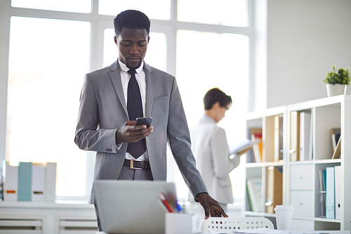 Young elegant African-american businessman in suit scrolling in smartphone while standing by workplace in office