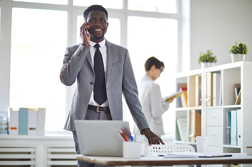 Happy young elegant businessman in suit talking on smartphone while standing by workplace in office