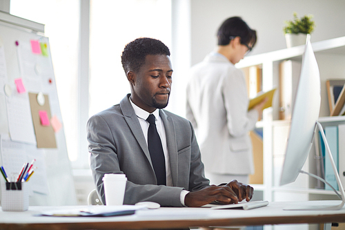 Young concentrated employee in formalwear typing on keypad in front of computer monitor during working day