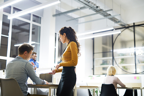 Serious attractive young lady mentor with curly hair giving papers and talking to young intern in modern office