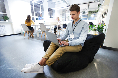 Serious concentrated handsome hipster manager with conference badge in neck sitting in bean bag and typing on laptop while analyzing data